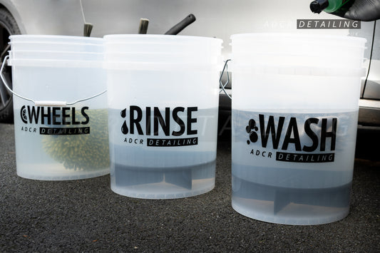 Three clear detailing buckets labelled 'Wheels', 'Rinse', and 'Wash' with decals and a car in the background.