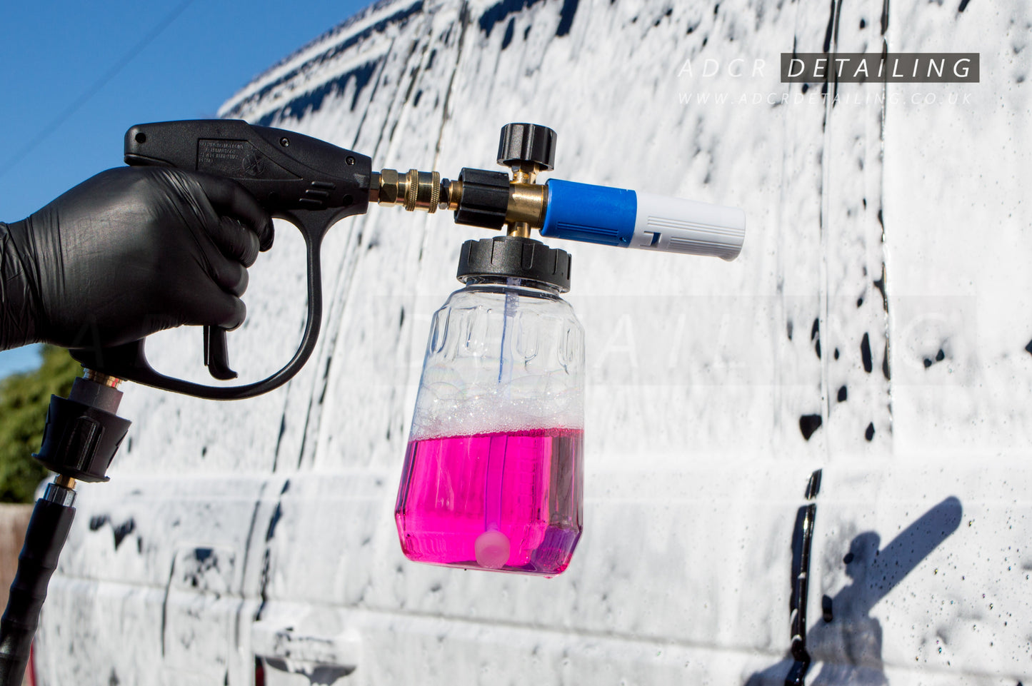 A hand holding a short trigger detailing gun with a clear V2 snow foam cannon filled with pink snow foam, against a white wall of snow foam on a car.