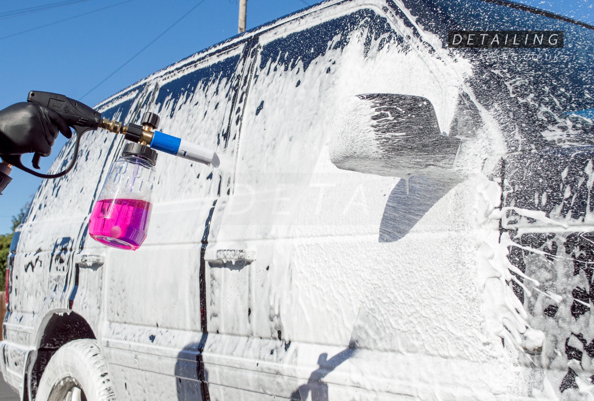 A hand holding a short trigger detailing gun with a clear V2 snow foam cannon filled with pink snow foam, spraying a thick white layer of snow foam on a car.
