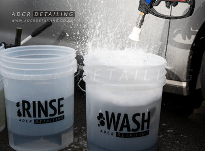 Two buckets labeled 'RINSE' and 'WASH' with a pressure washer in the background, used for car detailing.